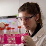 woman looking at test tubes, wearing goggles and white jacket