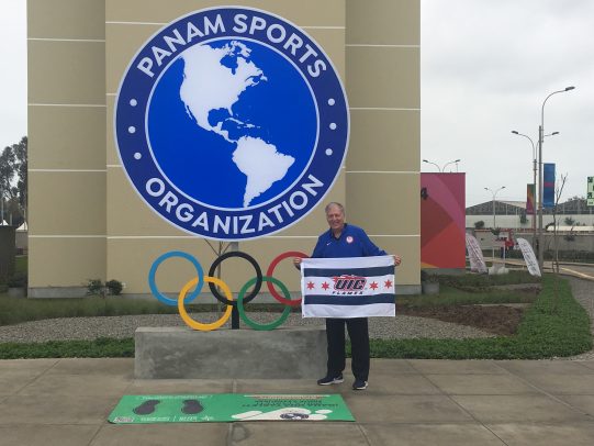 Dr. Mark Hutchinson holding a UIC Flames flag at the 2019 Pan American Games in Lima.