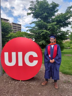 Anoop Nagabhushana, dressed in graduation attire, stands next to the UIC button sculpture in Memorial Grove.