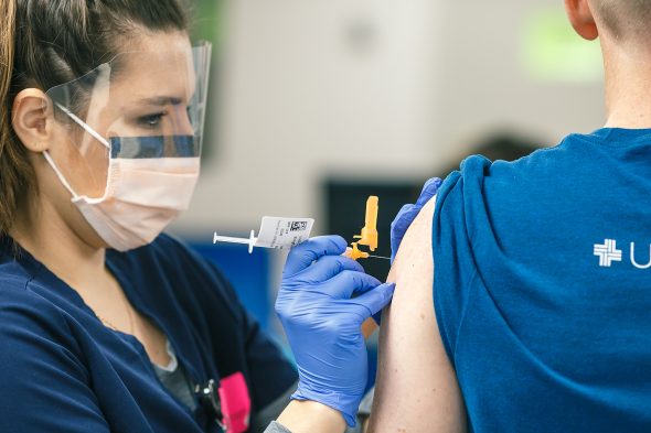 A woman wearing a face mask and a face shield puts a vaccine needle into a man's arm.