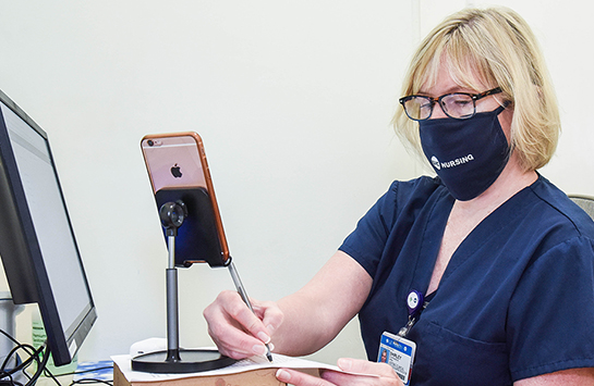 A female nurse wearing a UIC Nursing face mask and navy scrubs writes with a pen and paper and has an iPhone on a holder in front of her.