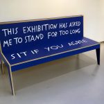 Alt text: Blue wooden bench in a gallery with text painted on it. The back of the bench reads, "This exhibition has asked me to stand for too long." The seat reads, "Sit if you agree."
