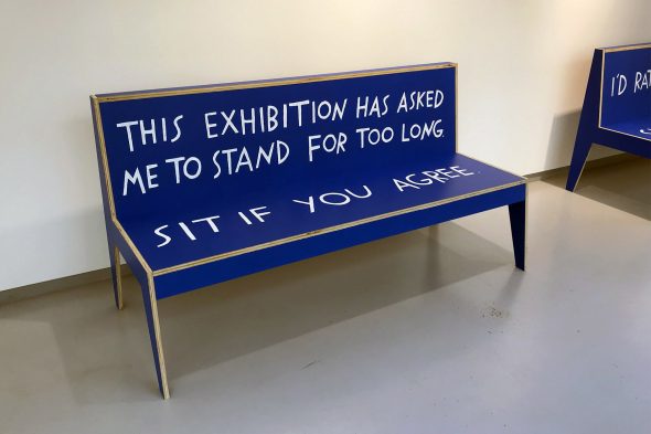 Blue wooden bench in a gallery with text painted on it. The back of the bench reads, "This exhibition has asked me to stand for too long." The seat reads, "Sit if you agree."