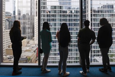 Five Break Through Tech Chicago Sprinterns (Wiktoria Zielinska, Chenille Lawrence, Jessica Alba, Nadia Taiym, and Trish Le) stand in a row with their backs to the camera and look at the city through large floor-to-ceiling windows with aluminum frames.