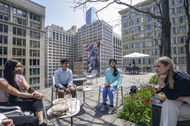 Three Break Through Tech Chicago Sprinterns (Jessica Alba, Nadia Taiym and Trish Le) sit on a rooftop in Chicago talking with a Morningstar employee (Anna Mukhina) after eating lunch. Mukhina is leaning in with her chin on her elbow and there are empty plates and glasses on the tables around them. There are multiple mid- and high-rise buildings around them.
