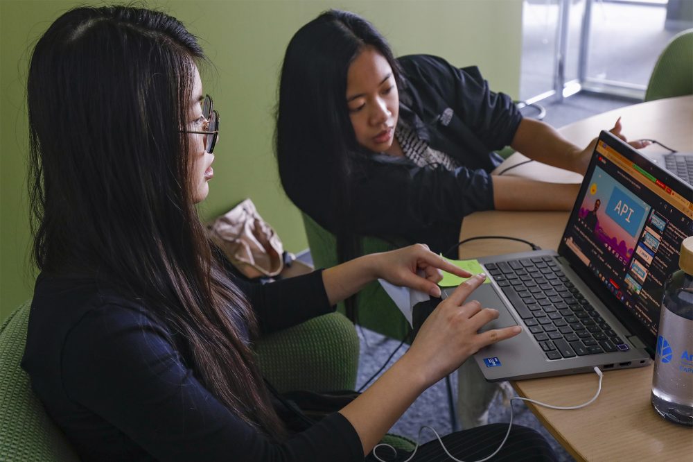 A UIC student (Trish Le) works at her computer in a Morningstar conference room while another UIC graduate (Jessica Alba) leans over and looks at the screen. They look focused.