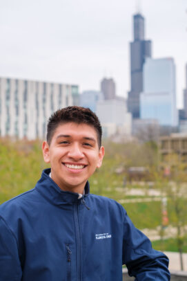 Christian Zavala wears a blue jacket and stands outside with the Willis Tower and Academic and Residential Complex in the background.
