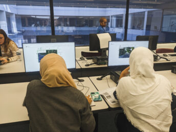 An image of two women at a computer lab sitting at a desk with two computers using the Project Sidewalk web page to rate google streetview imagery. The women have headscarves, with one being beige and one white.