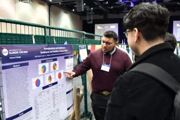 A man points at a research poster while a second man wearing a backpack listens.