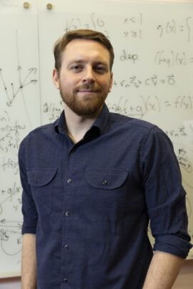A bearded man in a dark blue shirt stands in front of a white board filled with equations.