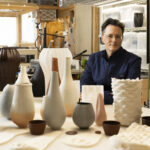 A man in a blue shirt and glasses sits behind several ceramic objects of various colors and shapes.
