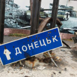 A street sign with a word in Ukrainian that is shot through with bullet holes lies on the ground.