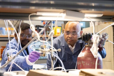 Two men in lab coats crouch to examine tubing in a laboratory.