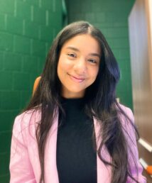 A woman with dark hair in a black shirt and pink blazer sitting in front of a green wall.