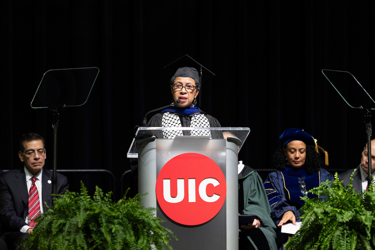 A woman in a graduation gown and cap speaks at a podium with a red circle and the letters UIC.