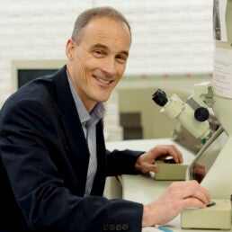 A man in a blazer sits at a microscope and smiles towards the camera.