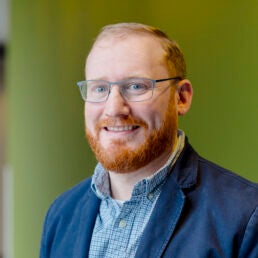 A man with red hair, a beard, and glasses poses against a green background.