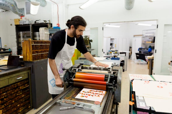 A man with a beard and glasses cranks a large metal machine in an print shop.