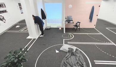 A room with a gray floor covered in tape marking the proposed location of walls and doors. A blanket and pillow are on the floor in the foreground. In the background, a man in black clothing leans out a window cut from a temporary interior wall.