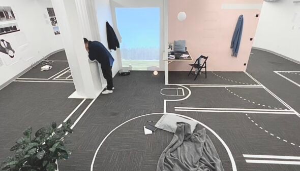 A room with a gray floor covered in tape marking the proposed location of walls and doors. A blanket and pillow are on the floor in the foreground. In the background, a man in black clothing leans out a window cut from a temporary interior wall.