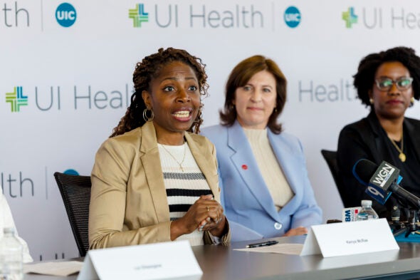 A seated woman speaks while two women look on in the background.