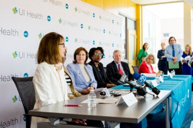 A group of seated men and women speak at news conference in front of a wall with the UI Health and UIC logos.