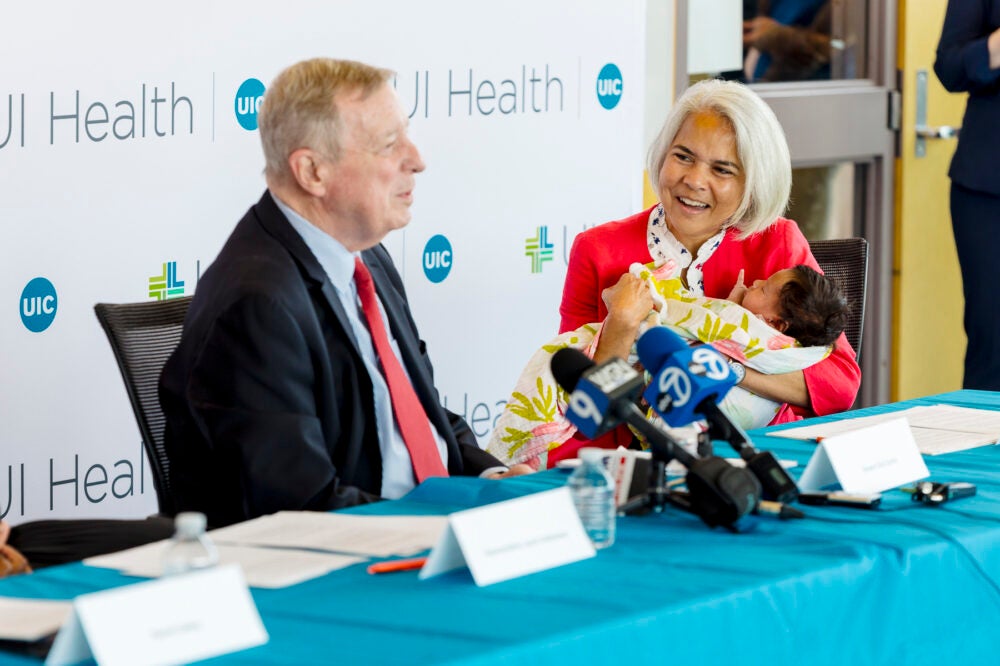 A seated man speaks in front of news microphones while a woman holding a baby watches and smiles.