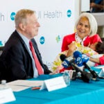A seated man speaks in front of news microphones while a woman holding a baby watches and smiles.