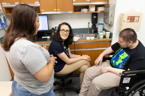 A doctor talks with a male patient in a wheelchair and woman standing nearby.