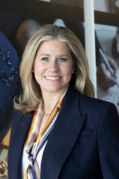 A woman in a navy blue blazer and a multi-colored scarf poses in front of a photo wall.