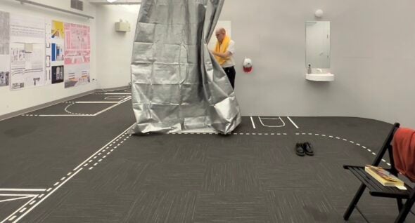 A room with a gray floor covered in tape marking the proposed location of walls and doors. At the back, a man with a yellow towel wrapped around his neck pulls back a silver curtain.