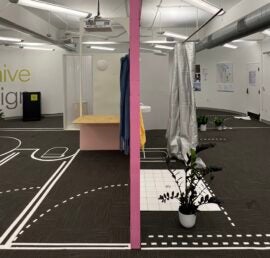 A pink wall vertically bisects two spaces in a gray-floored room with tape on the floor marking where walls and doors should go. In the left space sits a brow desk. On the right sits a potted plant, a rectangle of tiles, and a silver curtain.