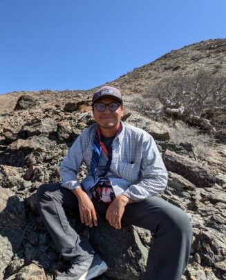 A man in a plaid shirt and baseball cap sits on a rock formation beneath blue skies.