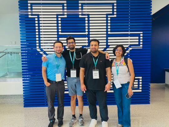 Three men and a woman wearing lanyards stand in front of a blue wall with the University of Kentucky logo.