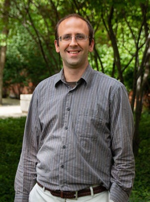 A man in a gray collared shirt stands outdoors in front of tress and greenery.