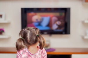 A little girl with pigtails seen from behind watching a flat-screen TV that is out of focus in the background.