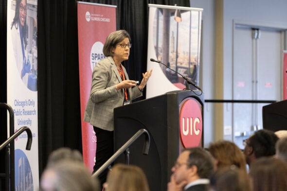 A woman in a gray blazer speaks at a podium with the red UIC logo on the front.