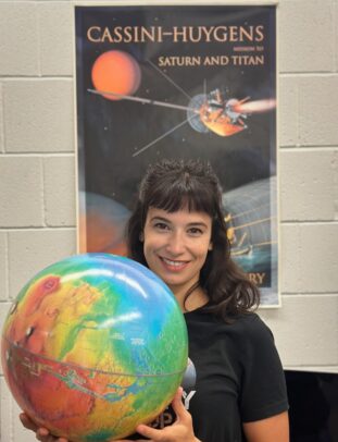 A woman with black hair holds a globe in front of a poster reading "Cassini-Huygens, Mission to Saturn and Titan" showing a satellite approaching a planet.