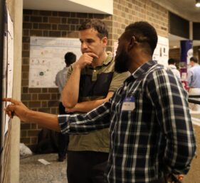 A man with his hand on his chin looks at a poster while a second man points at it.