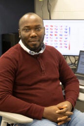 A man in a burgundy sweater sits in front o computer monitors showing climate maps of the United States.