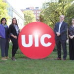 Three women and one man stand to either side of the red circle UIC logo on campus.