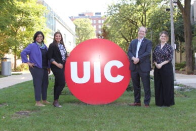 Three women and one man stand to either side of the red circle UIC logo on campus.