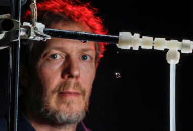 A slightly out-of-focus man stands behind black and white laboratory piping with a single drop of water falling.