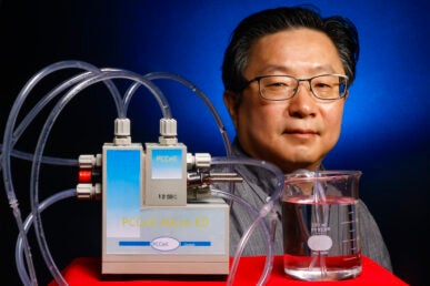 A man with glasses stands behind lab equipment with rubber tubing connected to a glass test tube filled with water.