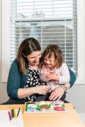 A woman in a blue cardigan holds a small child who is clapping her hands as both look at a puzzle.