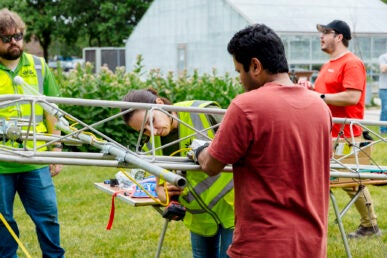 A woman in a neon green safety vest helps a man in a red shirt attach scientific instruments to a metal pole as another man in a neon green safety vest wearing sunglasses observes.