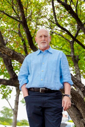 A man in a blue collared shirt stands outside in front of a tree.