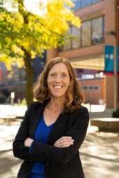 A woman in a black blazer and blue shirt stands outside in front of a brick building and a green tree.