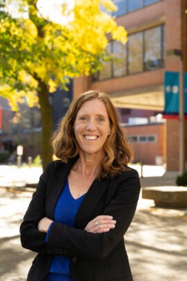 A woman in a black blazer and blue shirt stands outside in front of a brick building and a green tree.