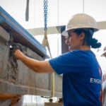 Two women wearing hard hats examine a cement and steel beam suspended from chains.